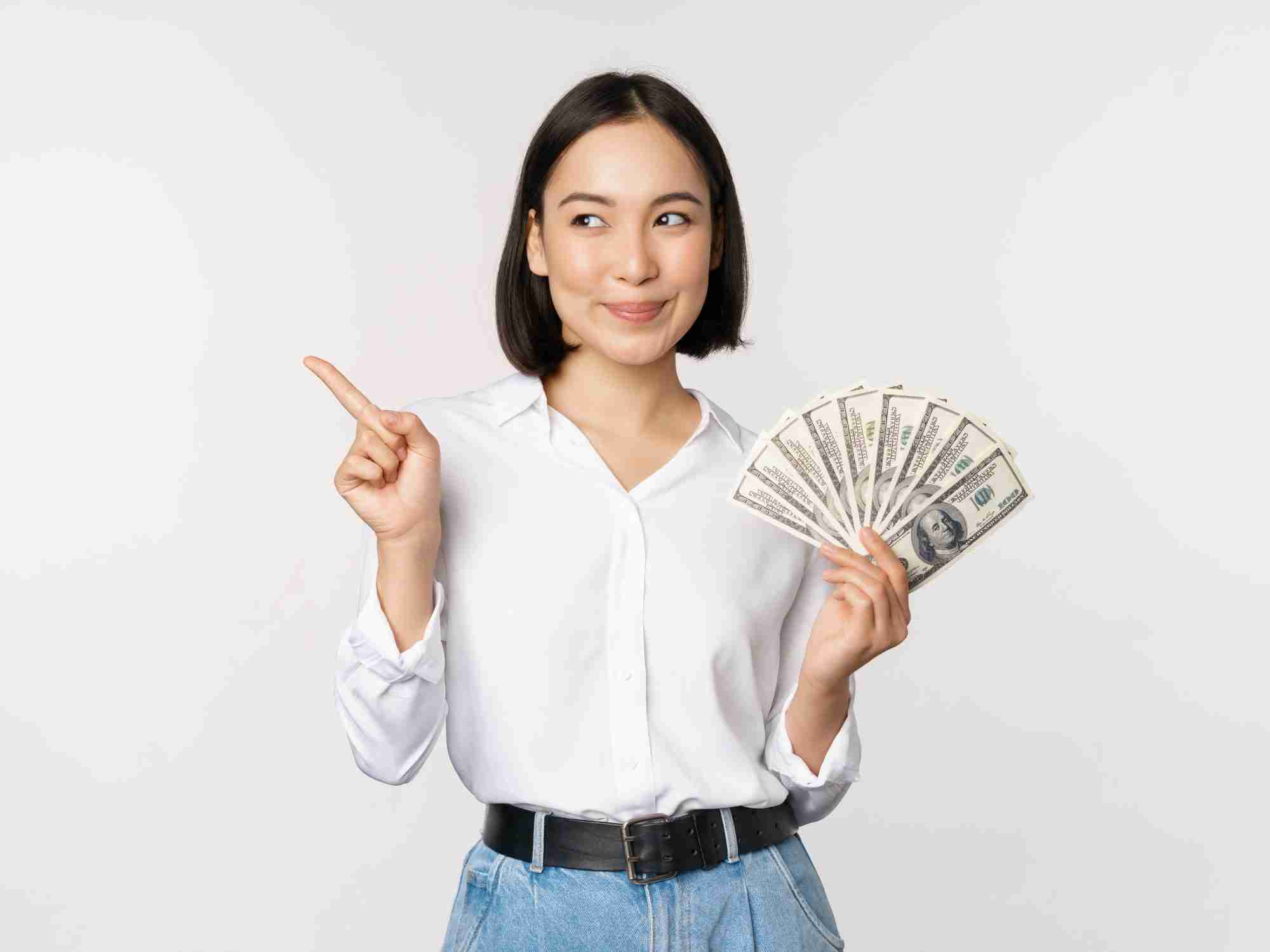 smiling young modern asian woman pointing banner advertisement holding cash money dollars standing white background edited scaled